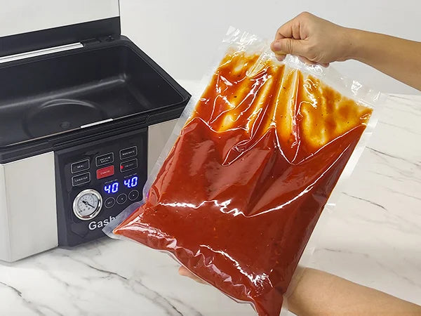 Hand holding a vacuum-sealed bag of food next to a vacuum sealer on a marble countertop.